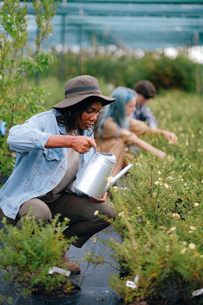 A woman tending plants in a greenhouse with a watering can, surrounded by greenery.